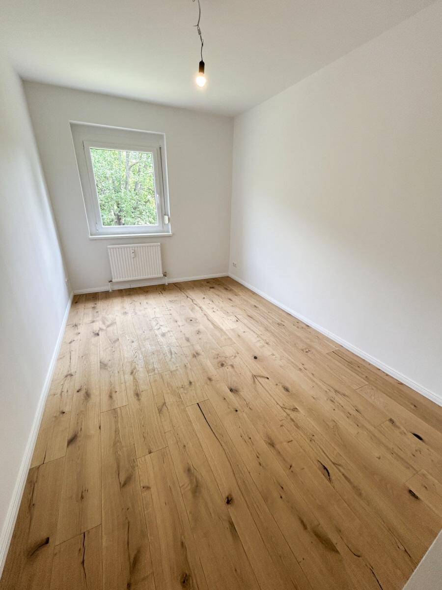 Kleineres Zimmer mit hellem Holzboden und einem Fenster mit Blick ins Grüne.