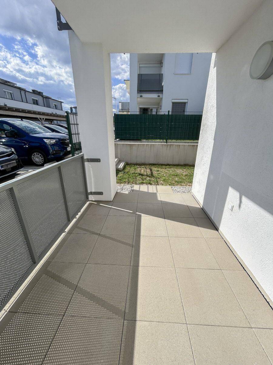 Geflieste Terrasse mit kleinem Gartenbereich und Blick auf den Parkplatz eines Neubaus.