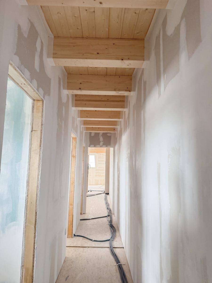 Langer Flur im Rohbau mit sichtbaren Holzbalken und vorbereiteten Wänden, führt zu weiteren Räumen.