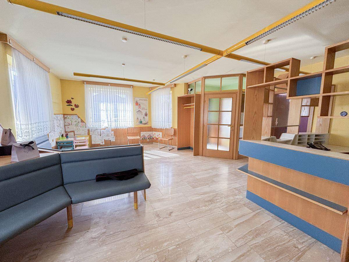 Heller Empfangsbereich mit großen Fenstern, Fliesenboden und einer Rezeptionstheke aus Holz.