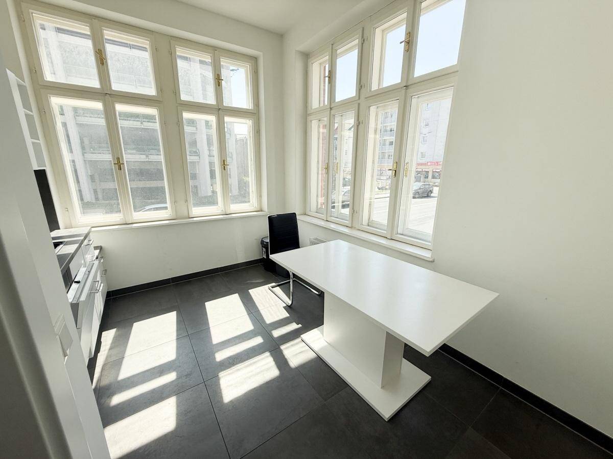 Kleiner, heller Raum mit dunklem Fliesenboden, großen Fenstern und einem weißen Tisch.