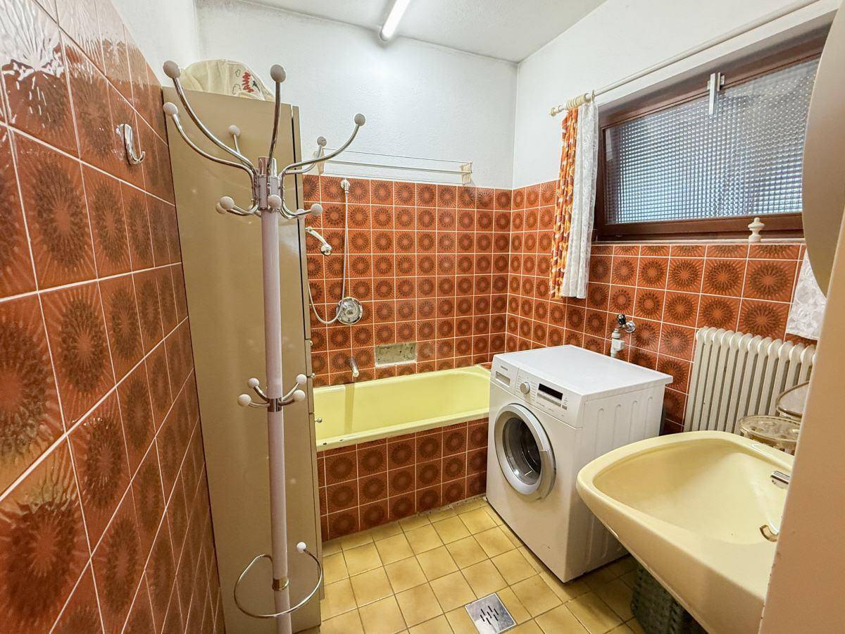 Badezimmer mit braunen Fliesen, Badewanne und Waschmaschine im 80er-Jahre-Stil.