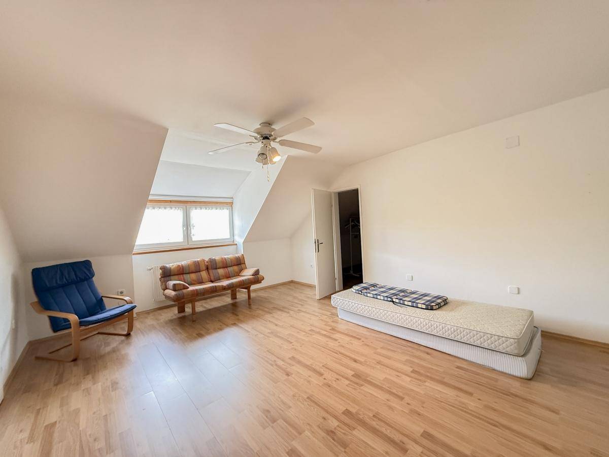 Geräumiges Dachzimmer mit schrägen Wänden, Holzboden und Deckenventilator.