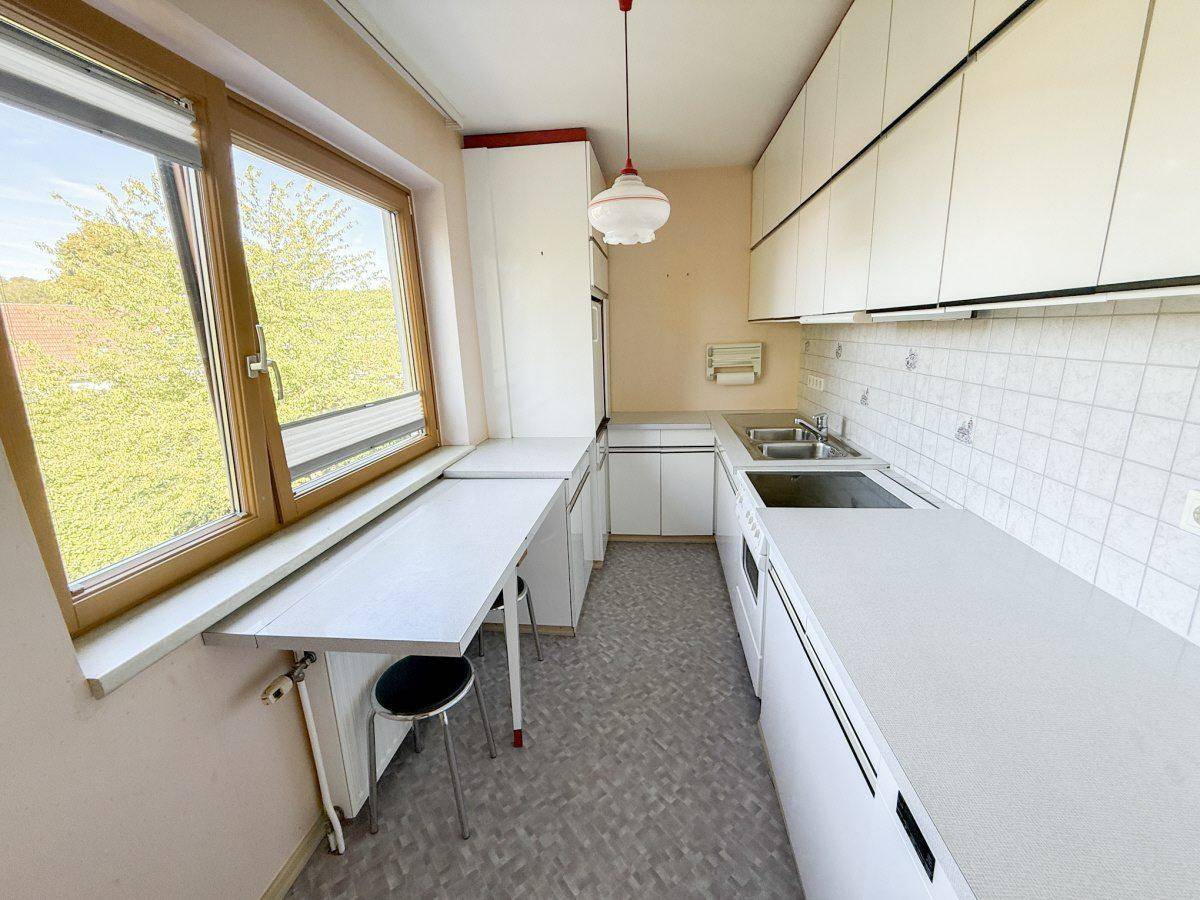 Ältere Küche mit weißen Schränken, integriertem Esstisch und großem Fenster mit Ausblick.