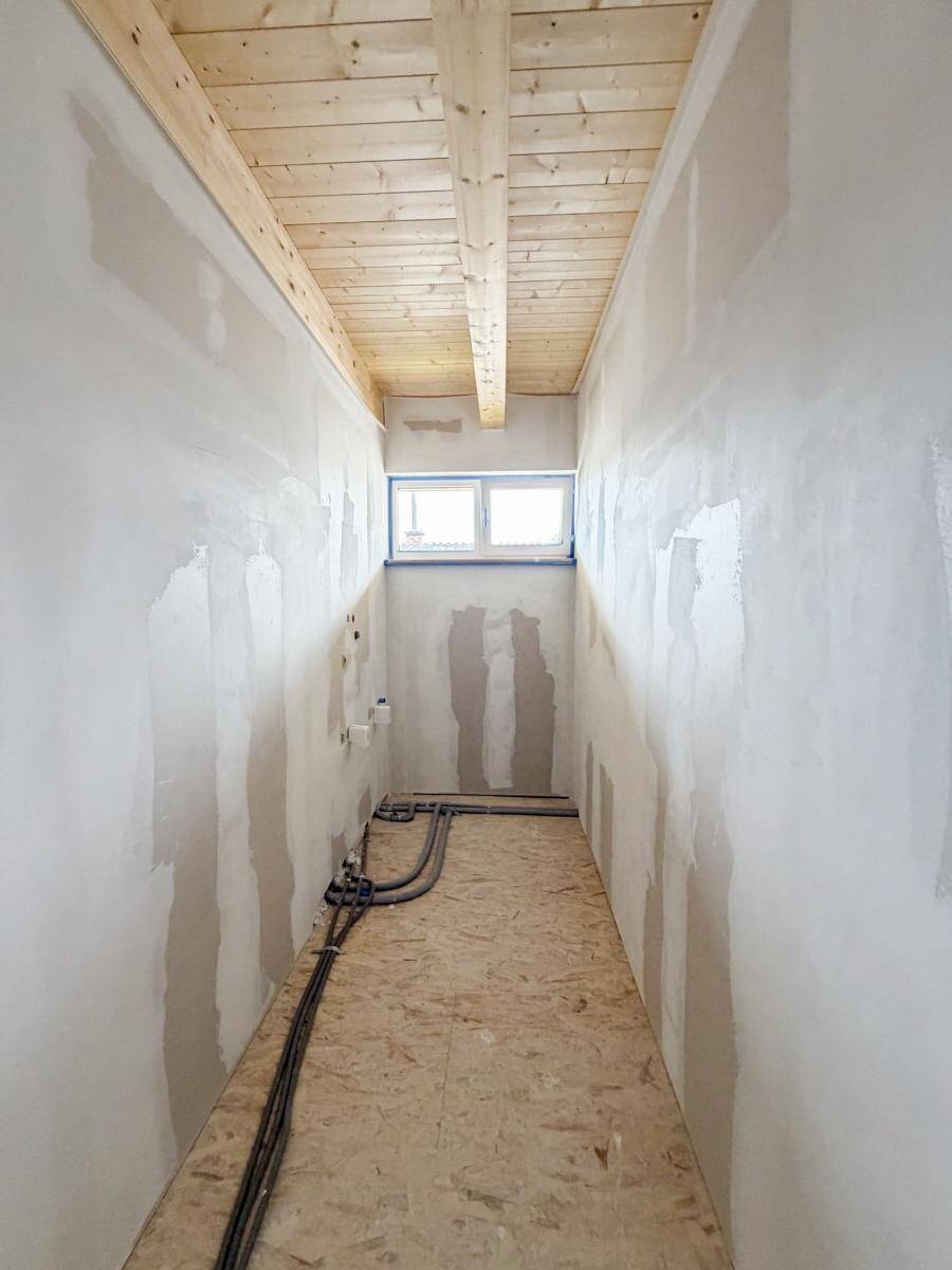 Schmaler Raum im Rohbau mit Holzbalkendecke, Fenster und vorbereiteten Sanitäranschlüssen.