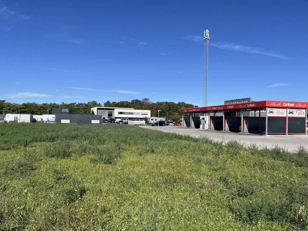 Grünes Feld mit Blick auf eine moderne Waschanlage und einen Mobilfunkmast unter strahlend blauem Himmel.