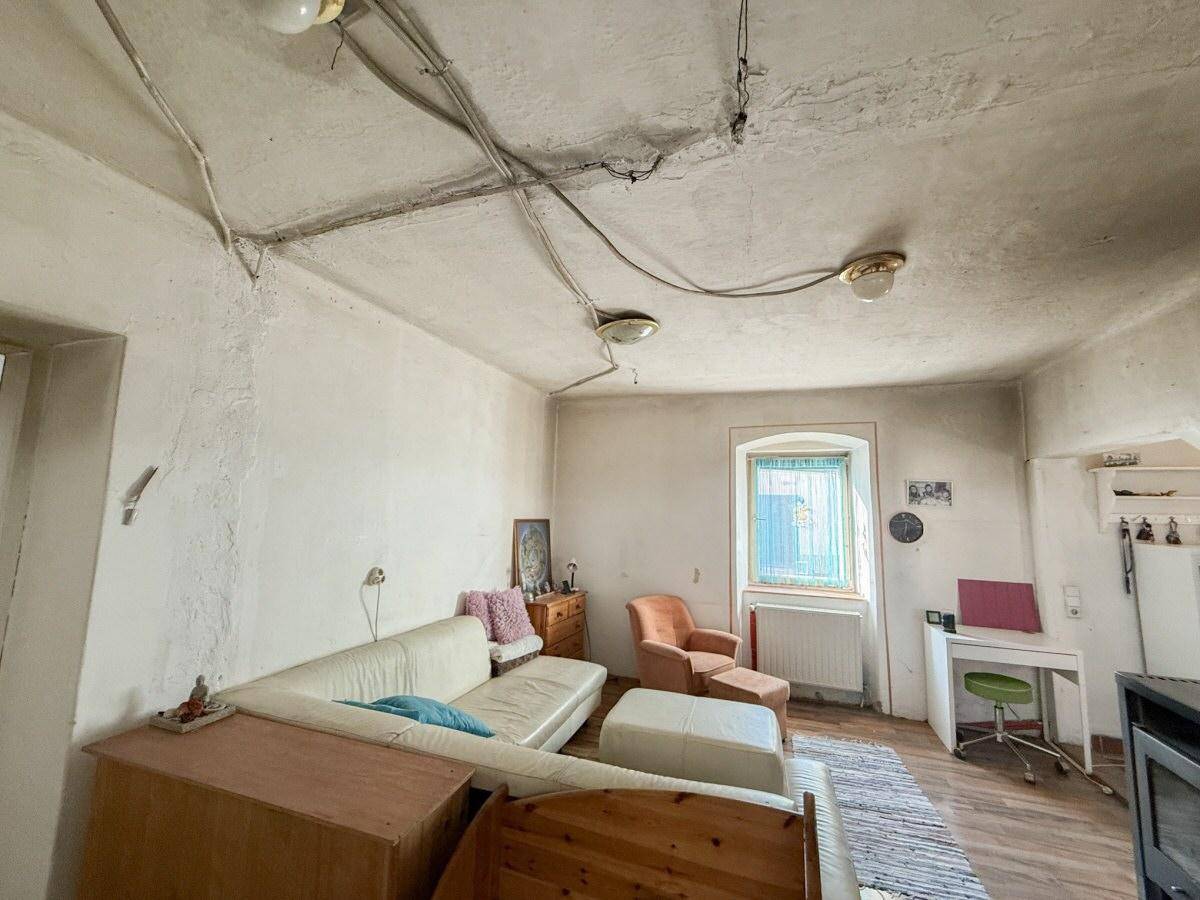 Wohnzimmer mit weiß gestrichenen Wänden, einem Sofa, Schreibtisch und sichtbaren Elektroleitungen an der Decke.