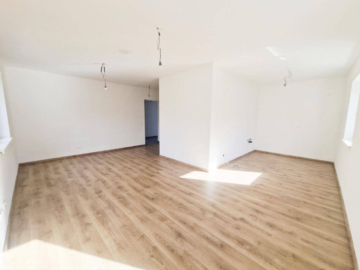 Geräumiger, leerer Wohnbereich mit hellem Laminatboden und viel natürlichem Lichteinfall durch die Fenster.