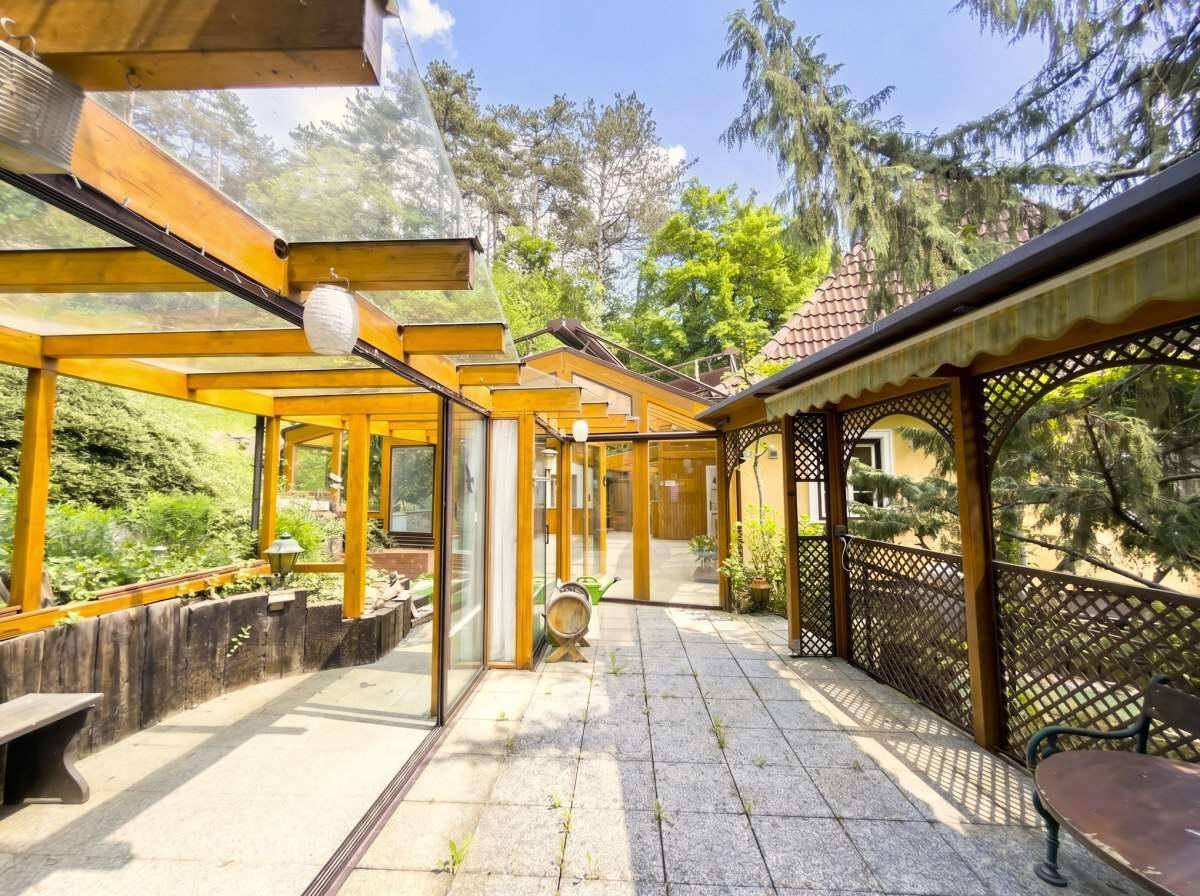 Große gepflasterte Terrasse mit Glasüberdachung und Holzelementen, umgeben von viel Grün.
