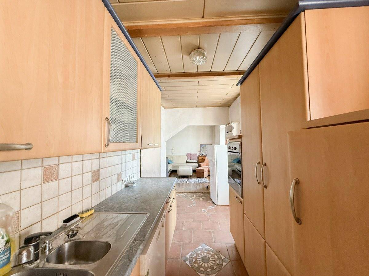Küche mit hellen Holzschränken, Spülbecken und Terrakottafliesenboden, mit Blick in einen weiteren Raum.