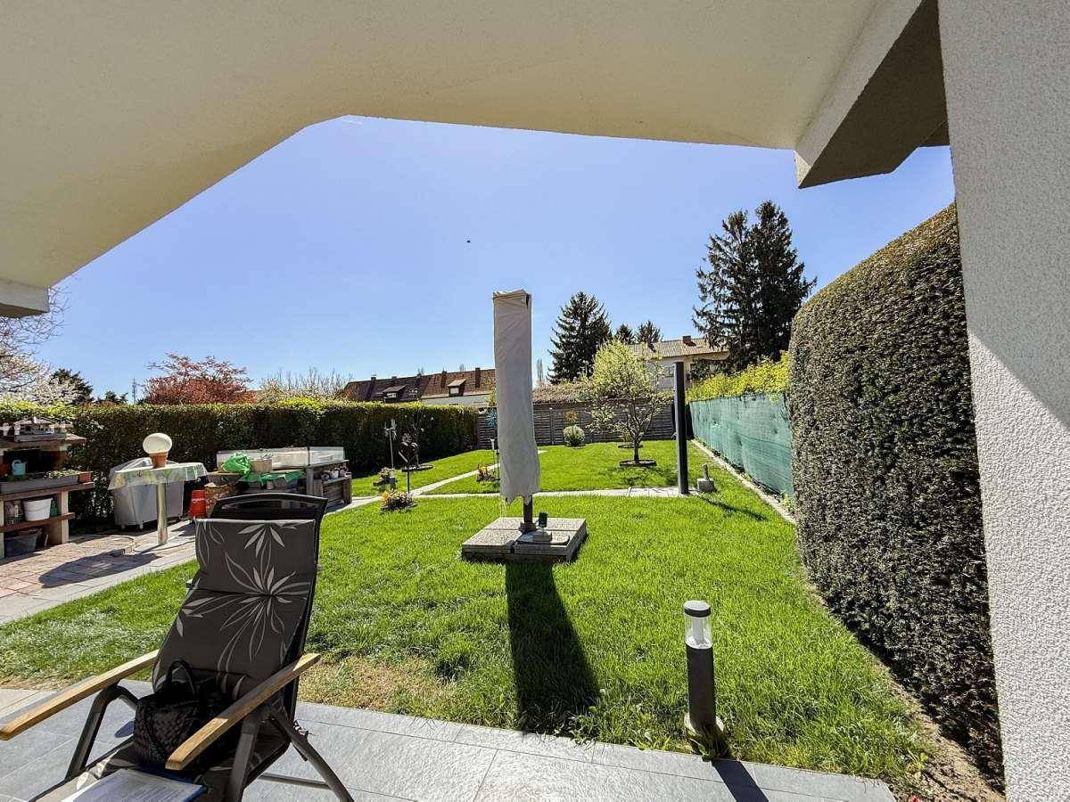 Blick von der überdachten Terrasse auf den grünen Garten mit hoher Hecke und viel Sonnenschein.