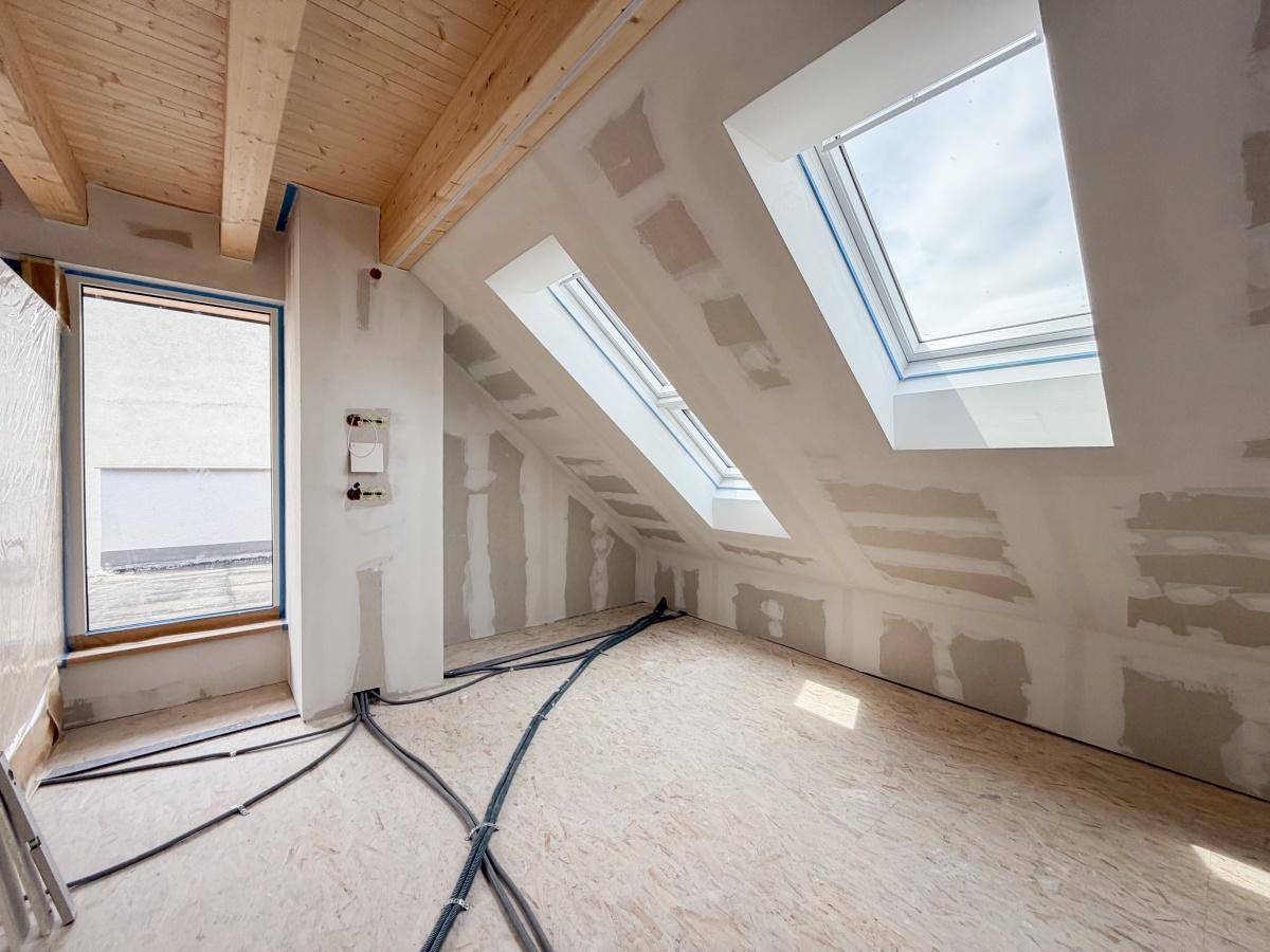 Rohbau eines Dachgeschosszimmers mit sichtbaren Holzbalken, Dachfenstern und Zugang nach draußen.