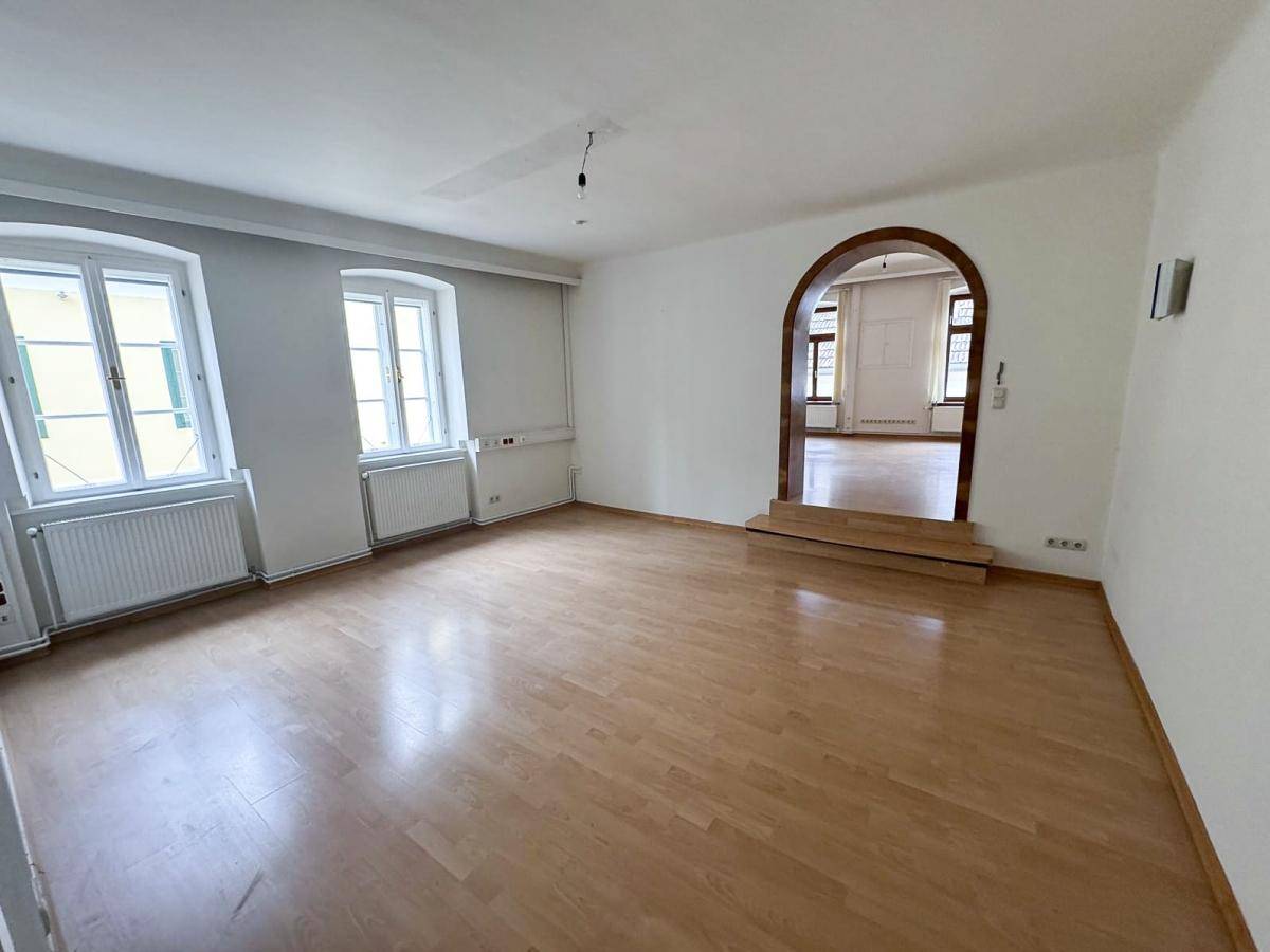 Geräumiges Wohnzimmer mit hellem Laminatboden, großen Fenstern und einem Rundbogen, der zu einem weiteren Raum führt.