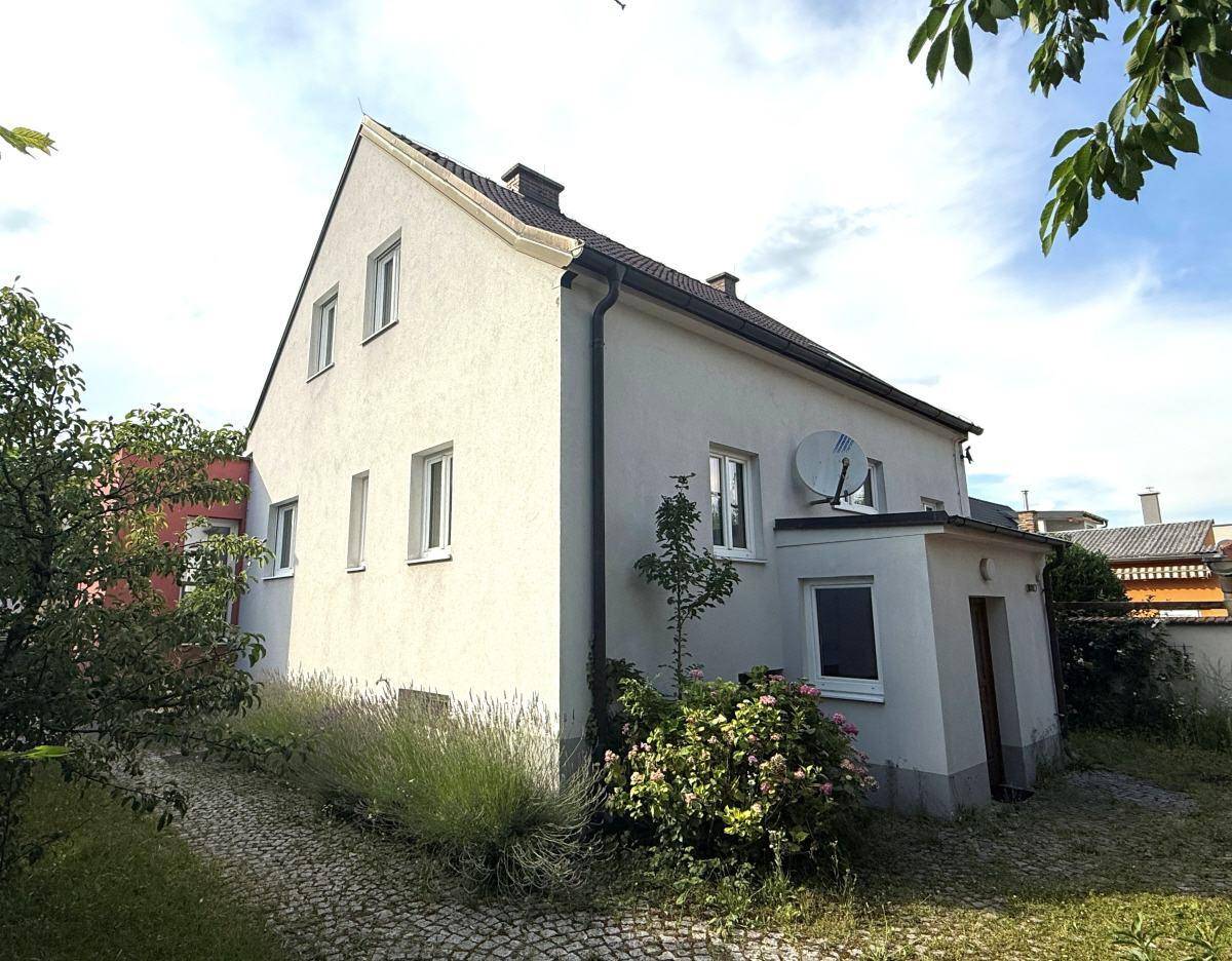 Seitenansicht des Hauses mit Garten, gepflastertem Weg und blühenden Pflanzen unter blauem Himmel.