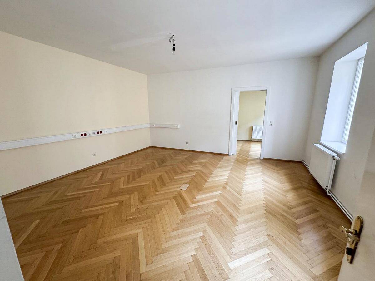 Großes Zimmer mit Fischgrätparkett, weißen Wänden und einer Fensterfront, die viel Licht spendet.