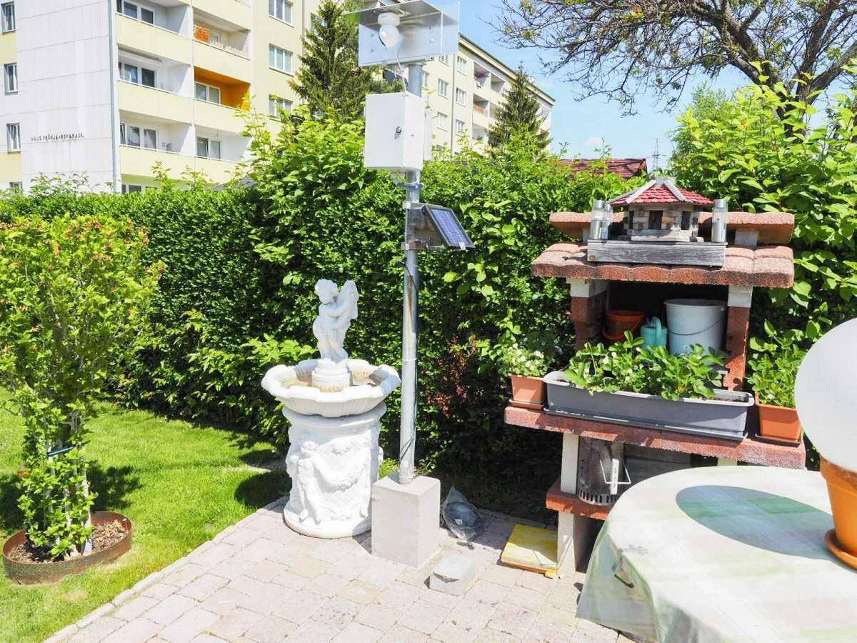 Dekorativer Gartenbereich mit einem Engelbrunnen und einer kleinen Gartenhütte unter blauem Himmel.