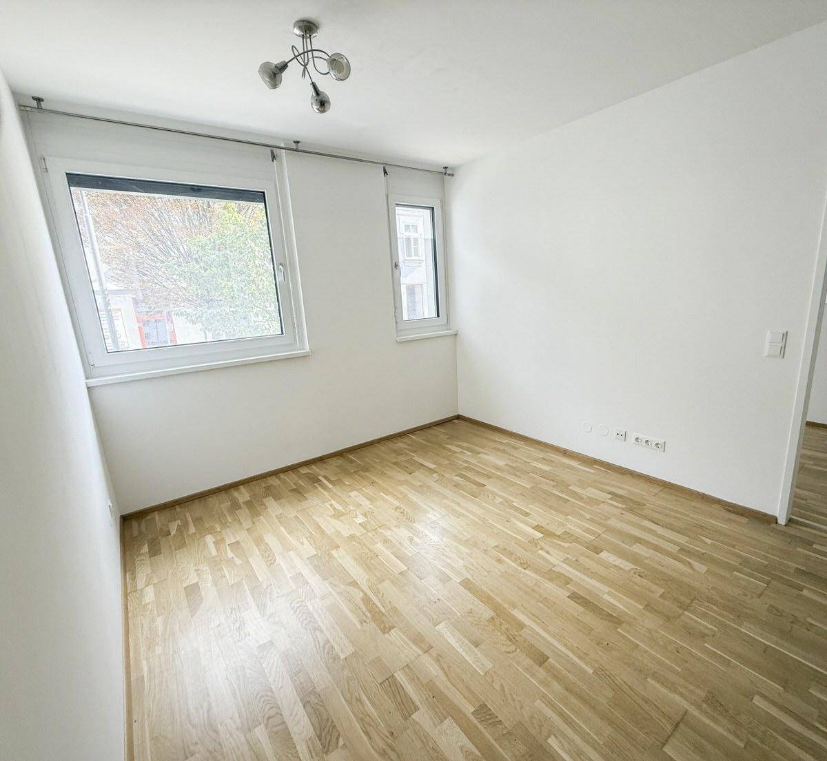 Helles Zimmer mit zwei Fenstern und hellem Holzparkett, ideal als Schlaf- oder Arbeitszimmer.