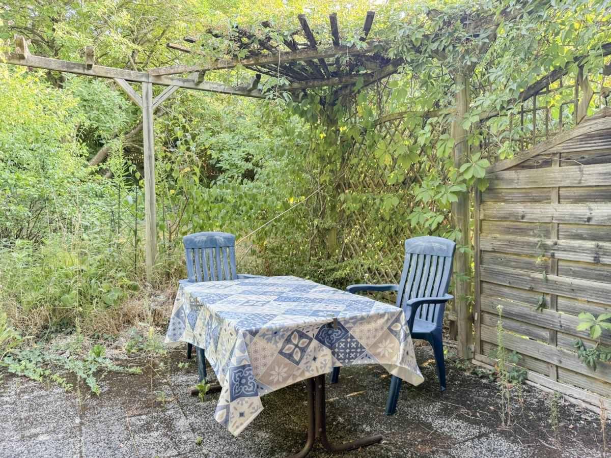 Gemütlicher Sitzbereich im Freien unter einer bewachsenen Holzpergola mit Tisch und Stühlen.