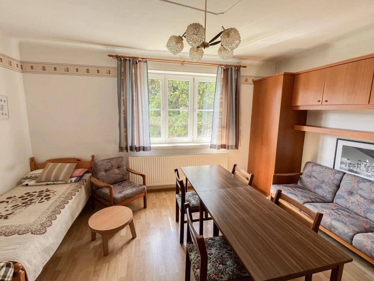 Schlafzimmer mit Einzelbett, Esstisch und Sofa, ausgestattet mit Holzmöbeln und einem Fenster.