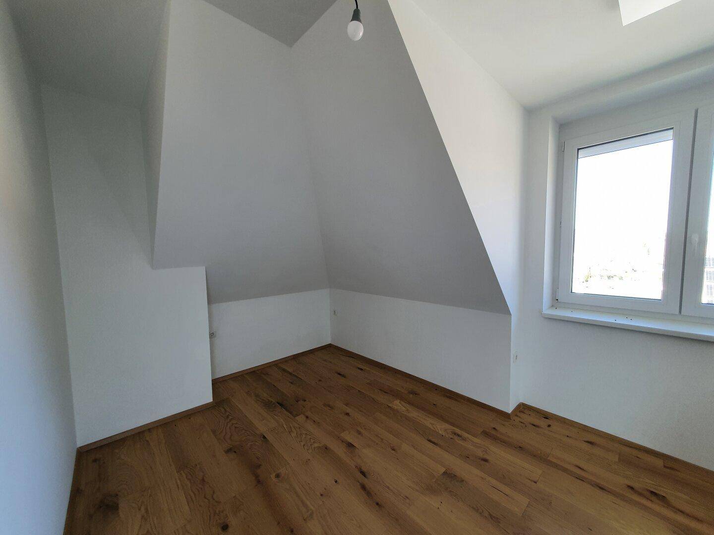 Gemütliches Zimmer im Dachgeschoss mit schrägen Wänden, Holzboden und Fenster.