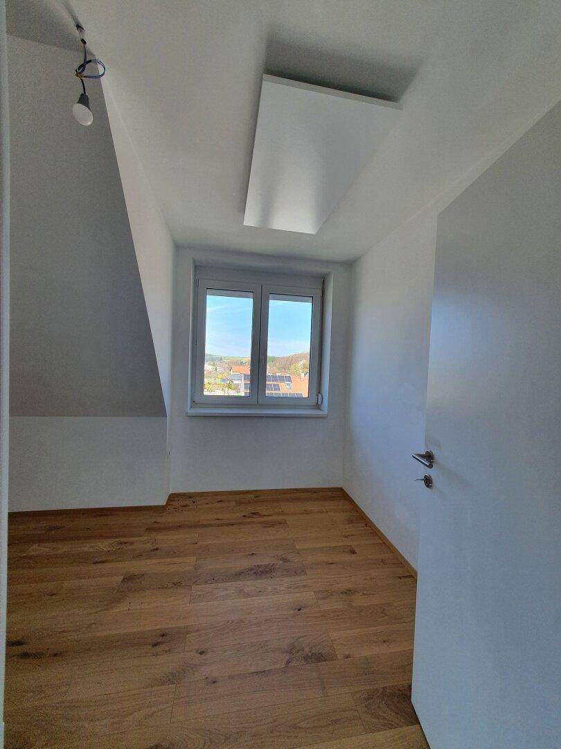Helles Dachzimmer mit Holzboden, schrägen Wänden und einem Fenster mit Ausblick.