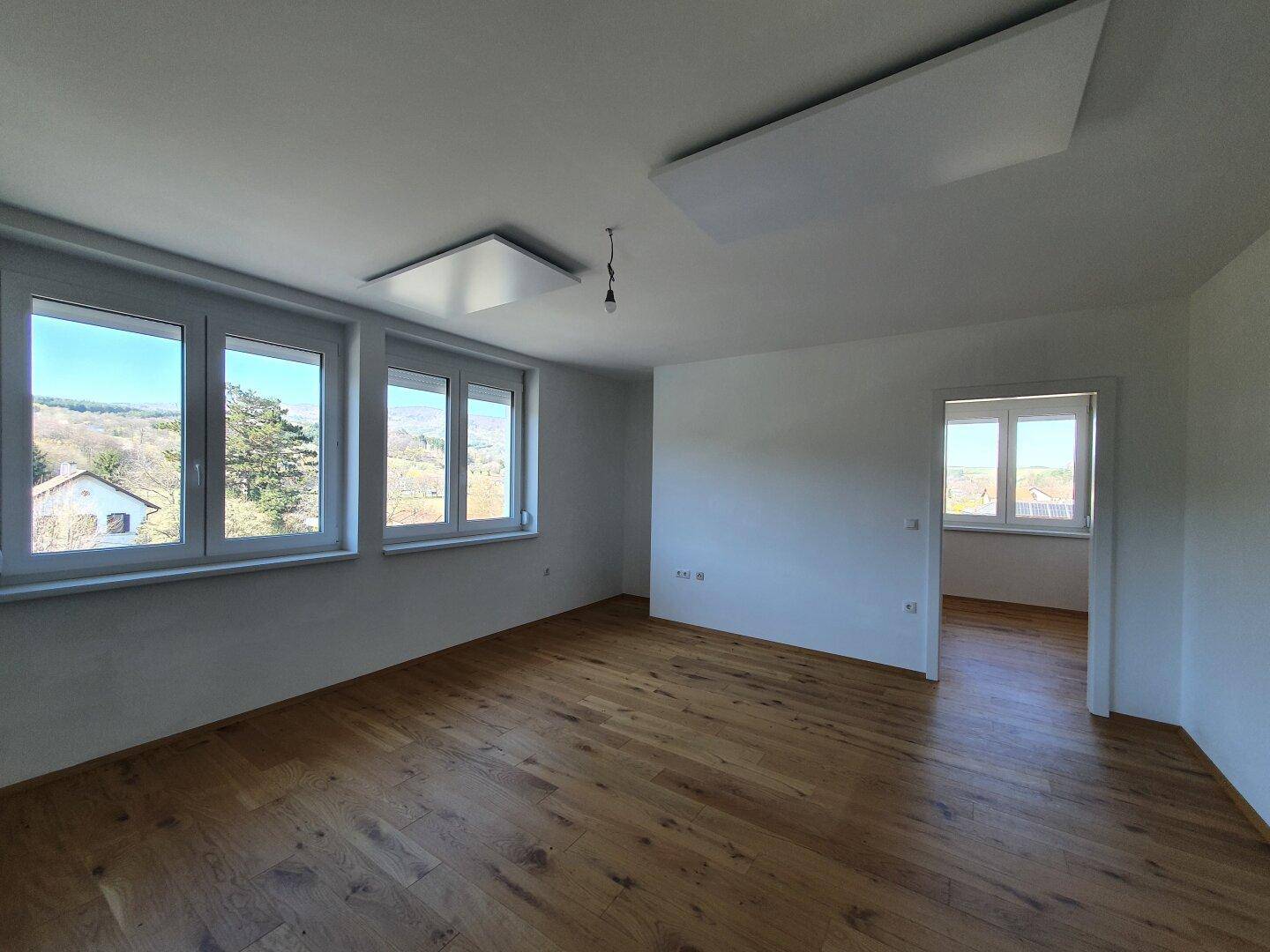 Geräumiger Wohnbereich mit Holzboden und großen Fenstern, die viel Licht hereinlassen.