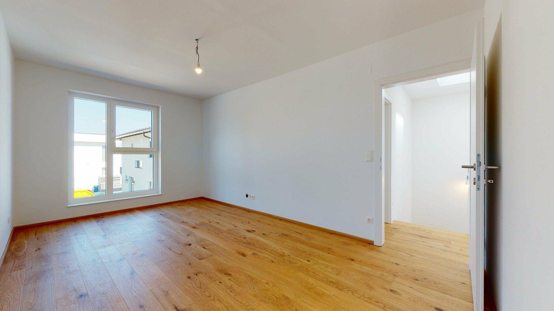 Geräumiger, heller Raum mit Holzboden und Fenster, bietet Zugang zu einem weiteren Bereich.