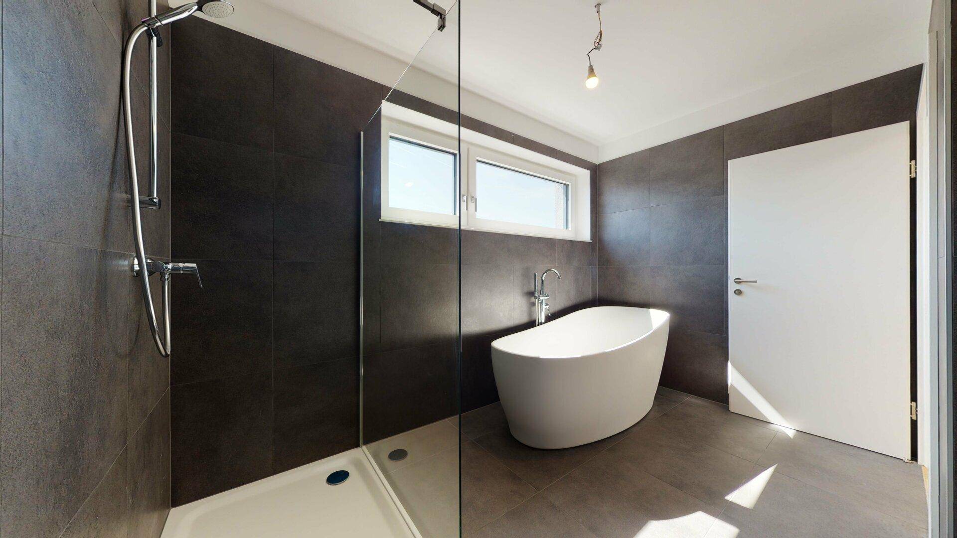 Modernes Badezimmer mit freistehender Badewanne, großer Dusche und dunklen Fliesen, hell durch Fenster.