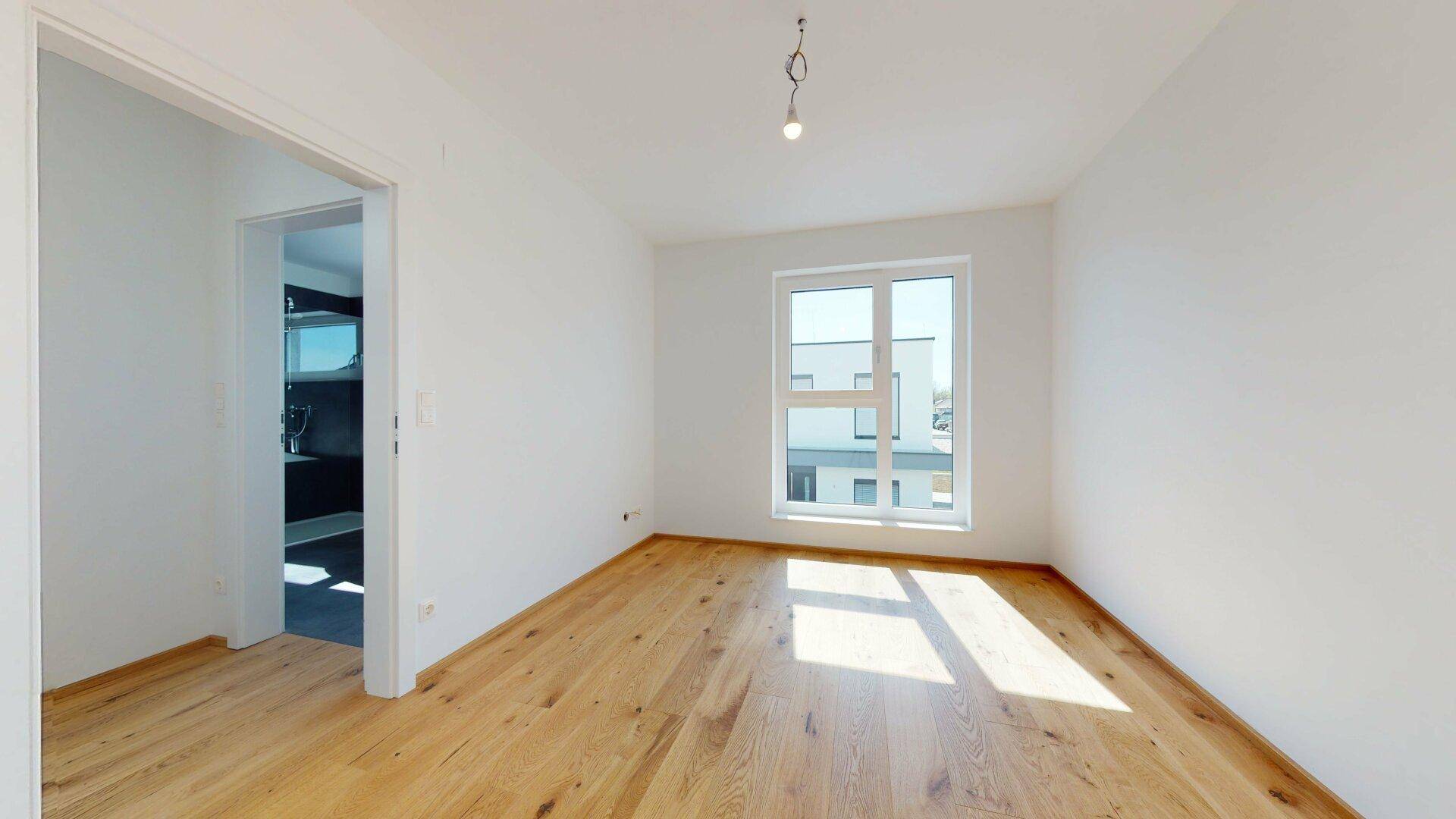 Heller Raum mit Holzboden und Fenster, bietet einen offenen Übergang zu einem angrenzenden Bereich.