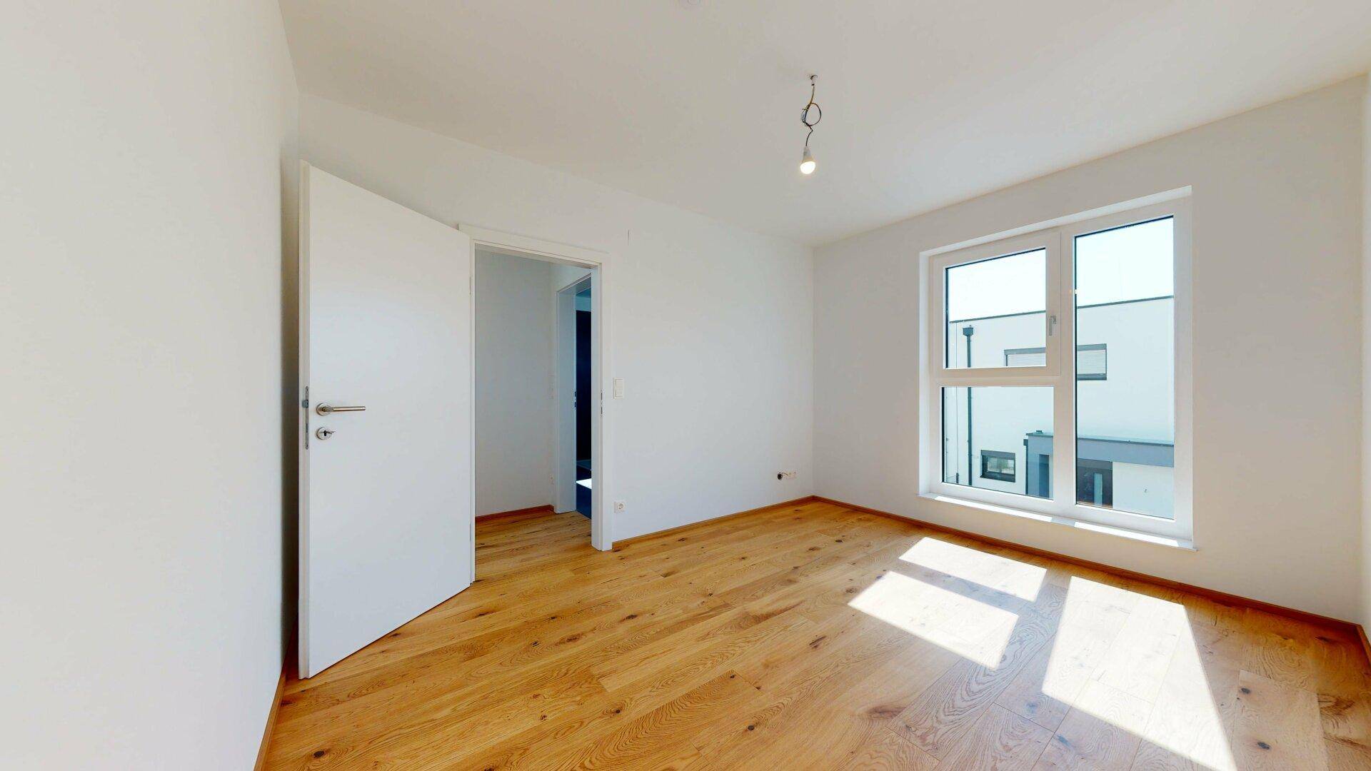Helles Zimmer mit Holzboden und Fenster, bietet viel Tageslicht und eine offene Tür.