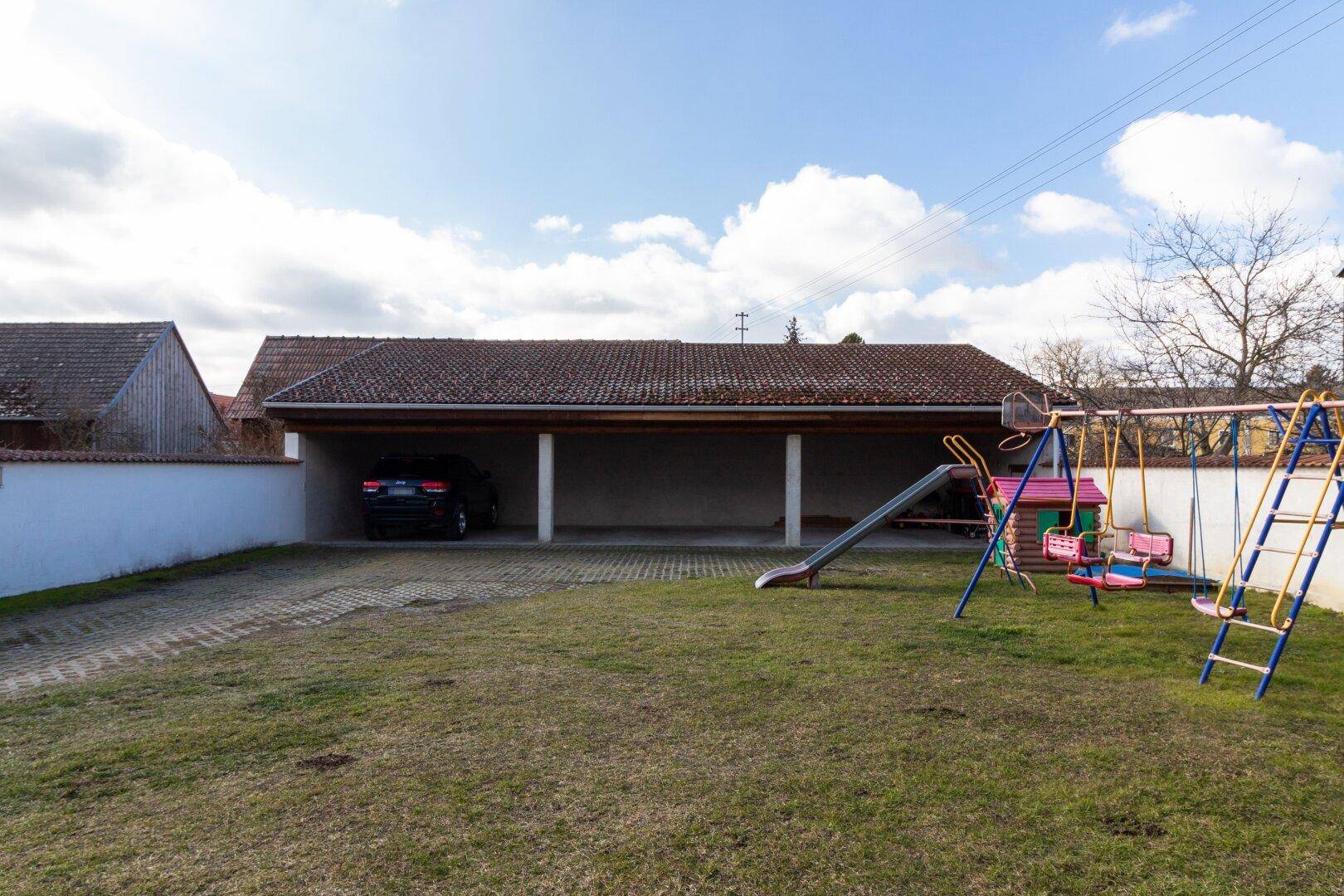 Hinterhof mit Kinderspielplatz, Rasenfläche und einem überdachten Carport für Fahrzeuge.