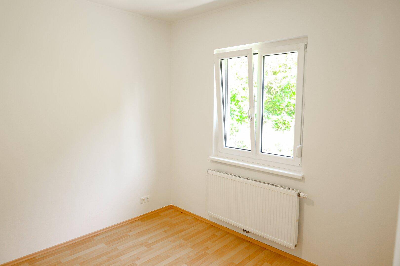 Leeres Zimmer mit Laminatboden, weiß gestrichenen Wänden und Fenster mit Blick ins Grüne.