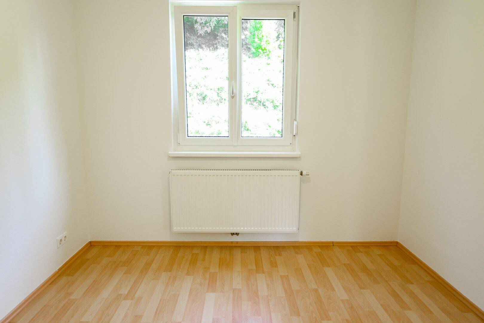 Leeres Zimmer mit Laminatboden, großem Fenster und Heizkörper unter dem Fenster.