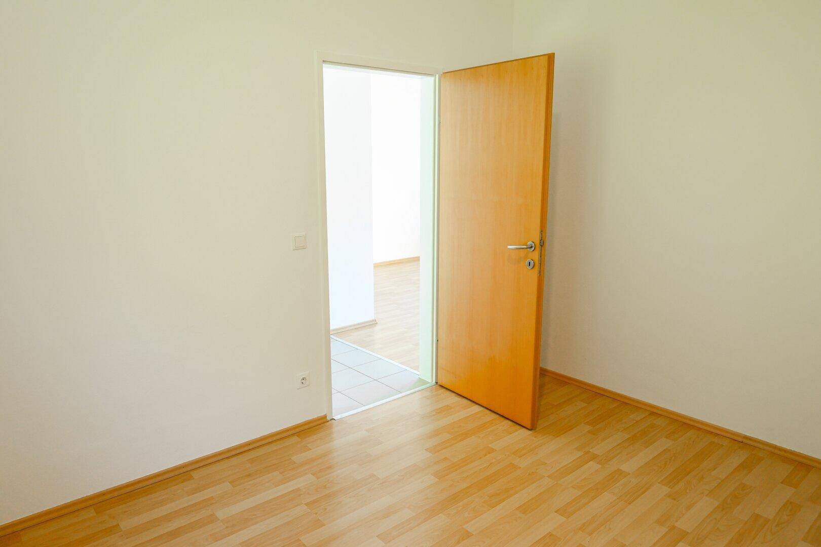 Leeres Zimmer mit Laminatboden und offener Holztür, die in einen anderen Raum führt.