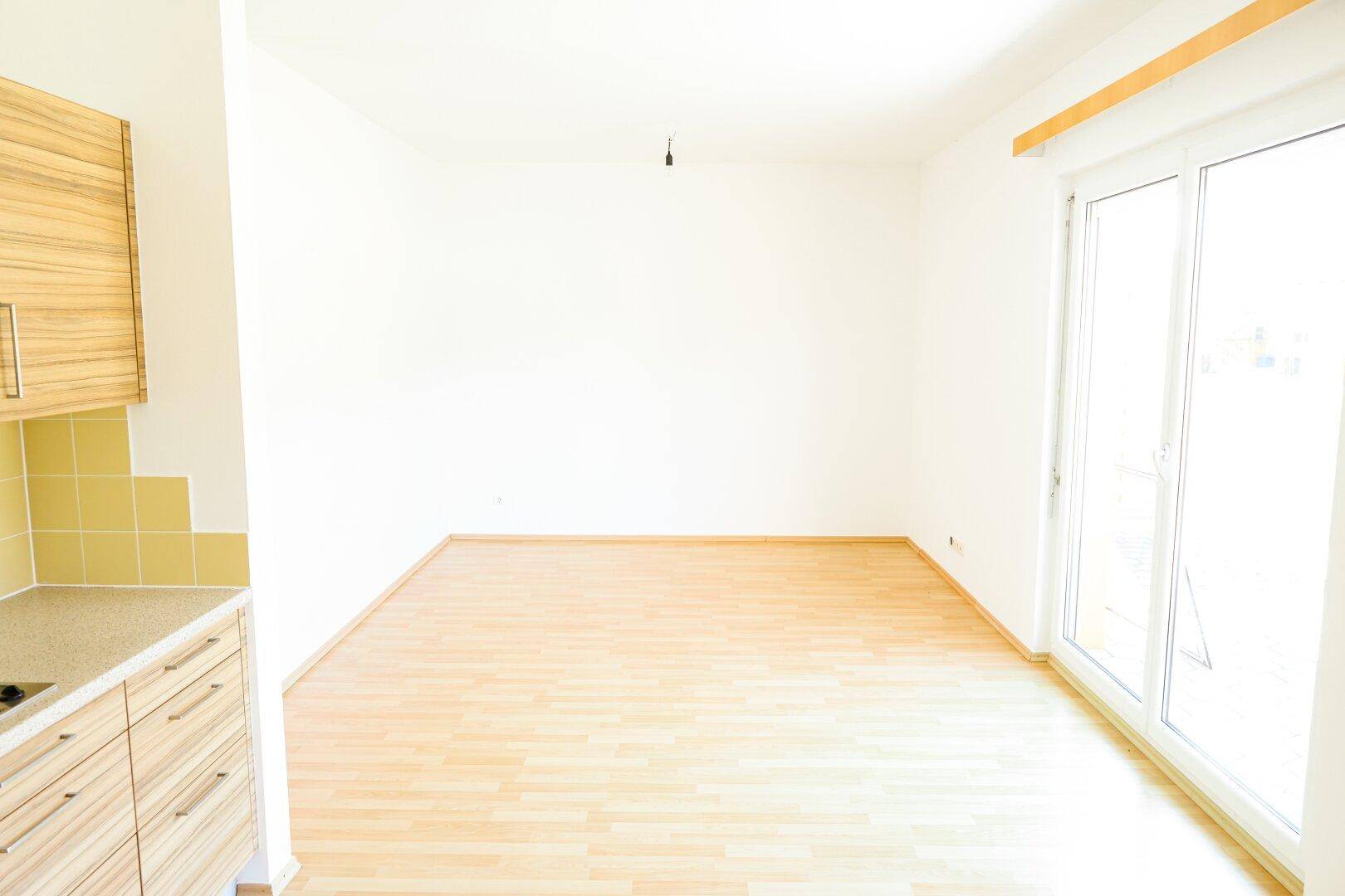 Heller, leerer Raum mit Laminatboden und angrenzender Küchenzeile.