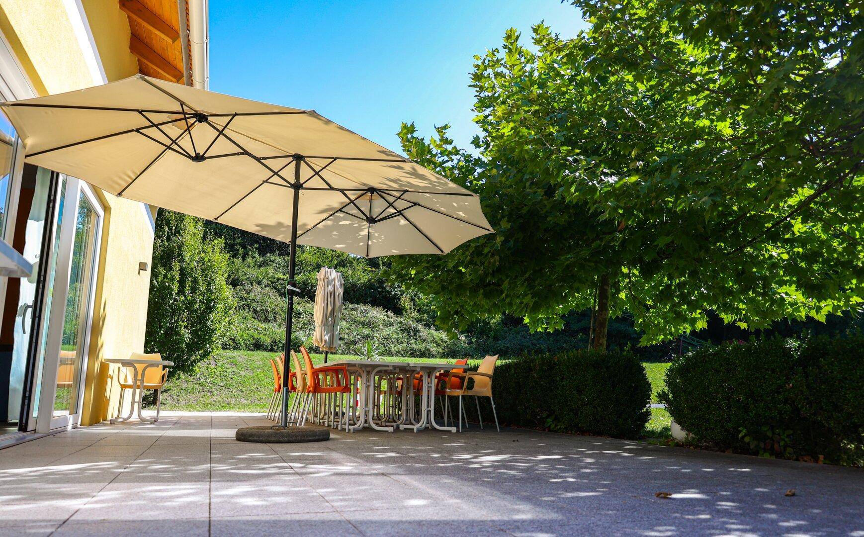 Große gepflasterte Terrasse mit Sitzgelegenheiten und Sonnenschirm unter Bäumen.