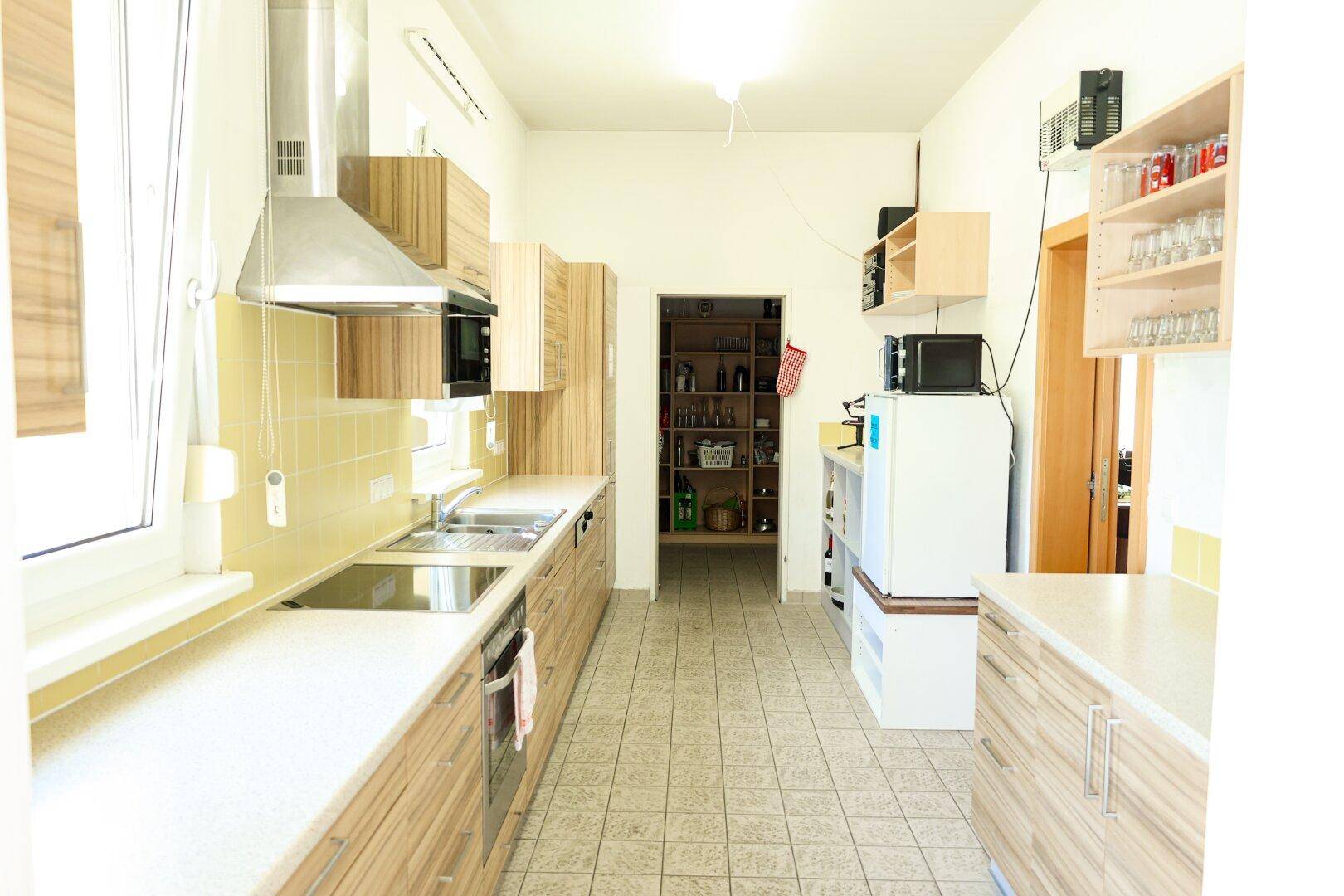 Lange Küche mit Fliesenboden, Holzschränken und Durchgang zu einer Speisekammer.