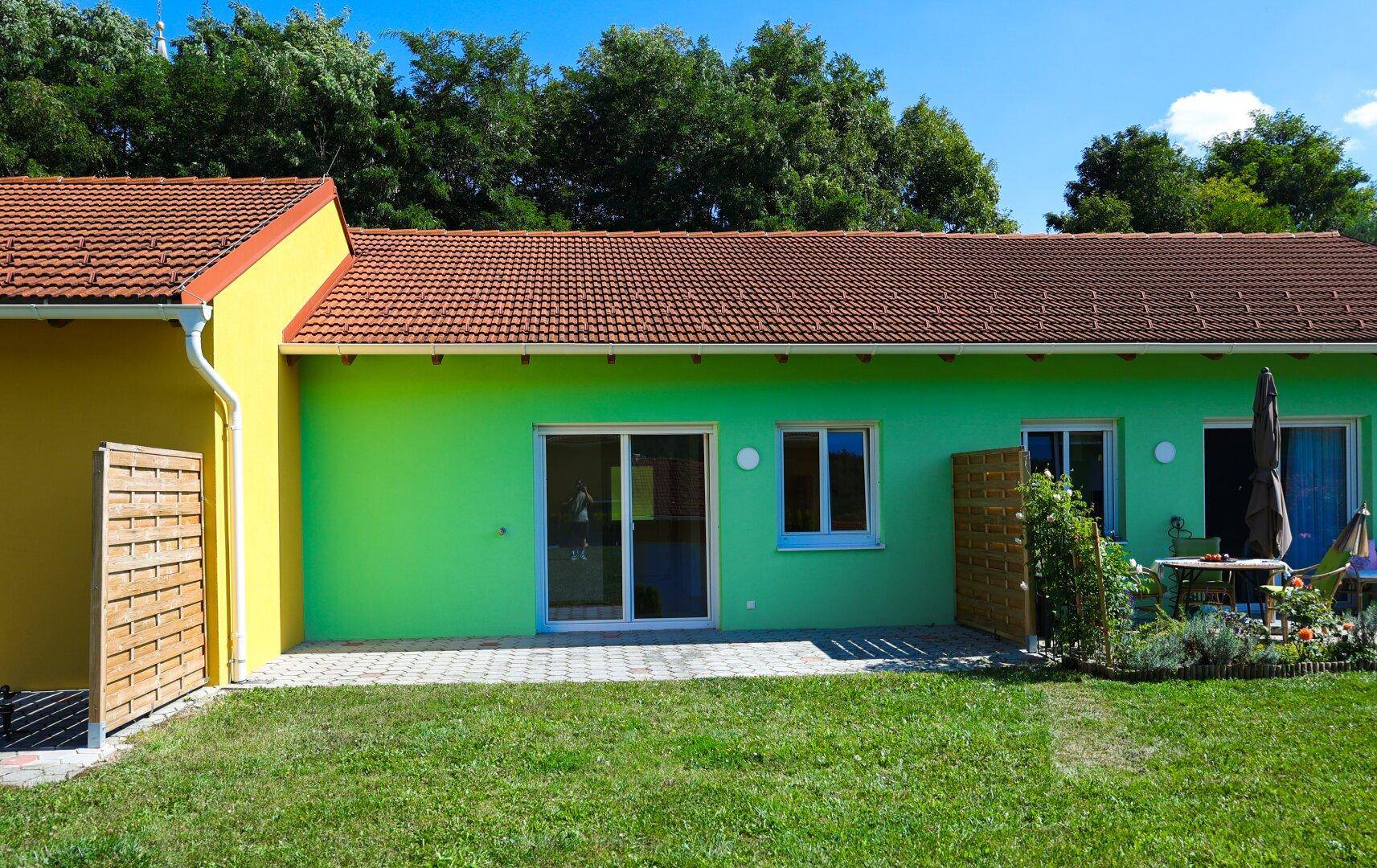 Grünes Haus mit gepflasterter Terrasse, Sitzgelegenheiten und Gartenbereich.