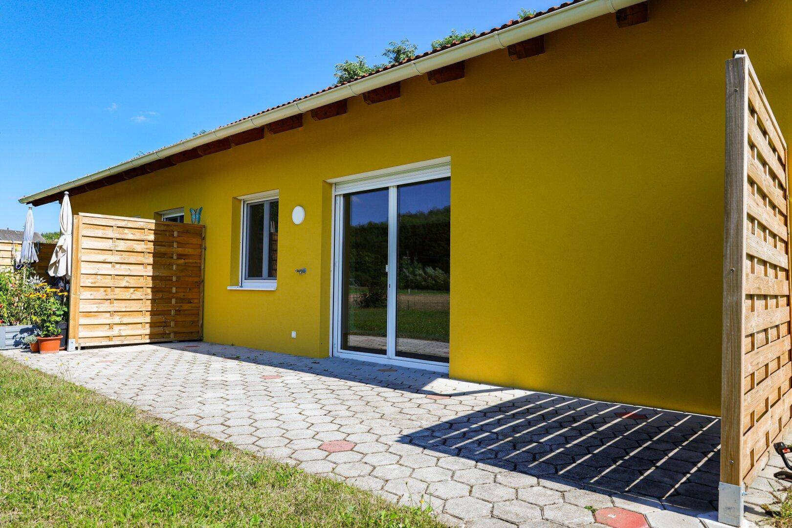 Gelbes Haus mit gepflasterter Terrasse und hölzernen Sichtschutzelementen.