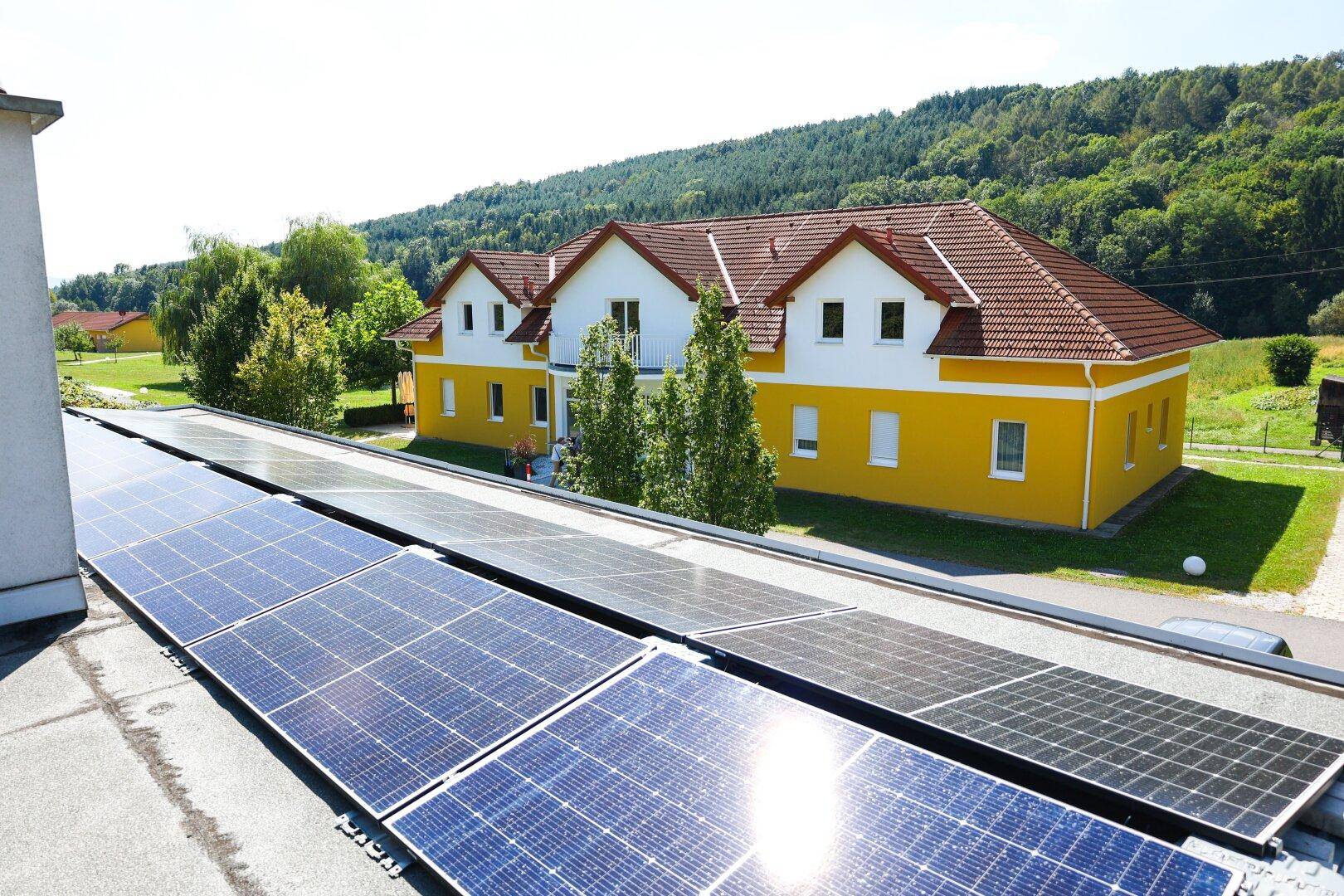 Photovoltaikanlage auf einem Dach mit einem gelben Gebäude und grüner Landschaft im Hintergrund.