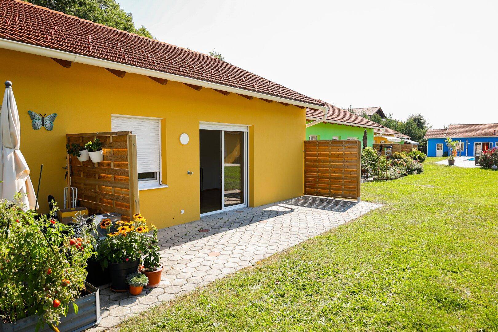 Gelbes Haus mit kleiner gepflasterter Terrasse und Gartenbereich unter blauem Himmel.