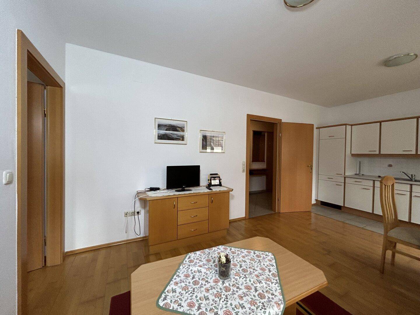 Wohnbereich mit Holzboden, Fernsehmöbel und Zugang zur Küche und weiteren Räumen.