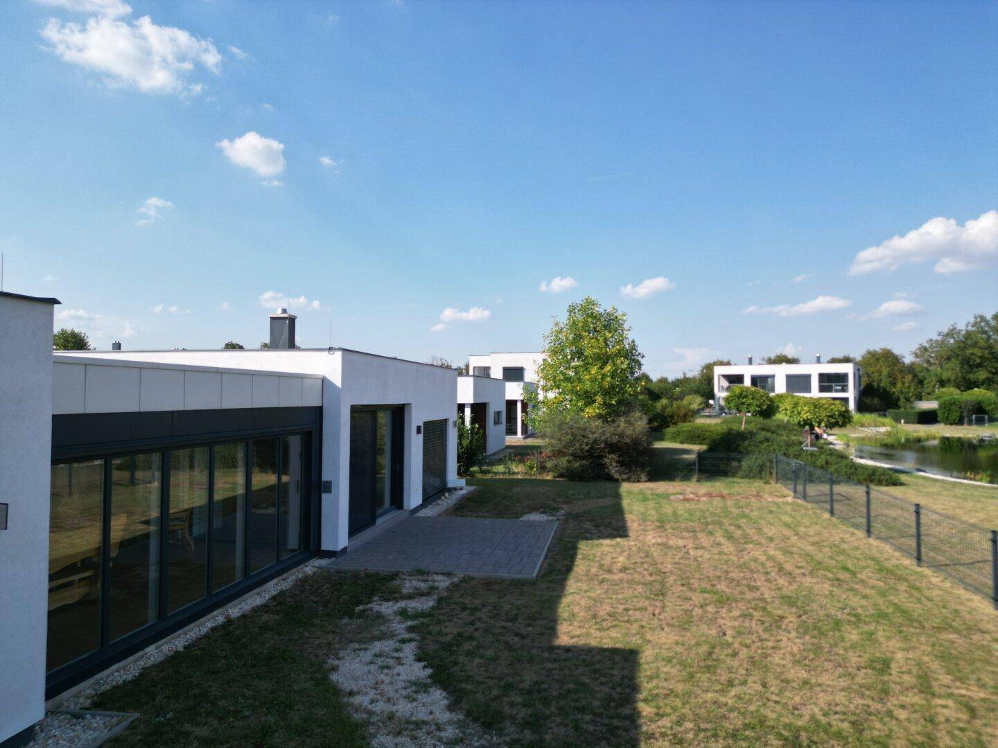 Hinterhof eines modernen Hauses mit einer gepflasterten Terrasse und einem Rasen, der zu einem Teich führt.