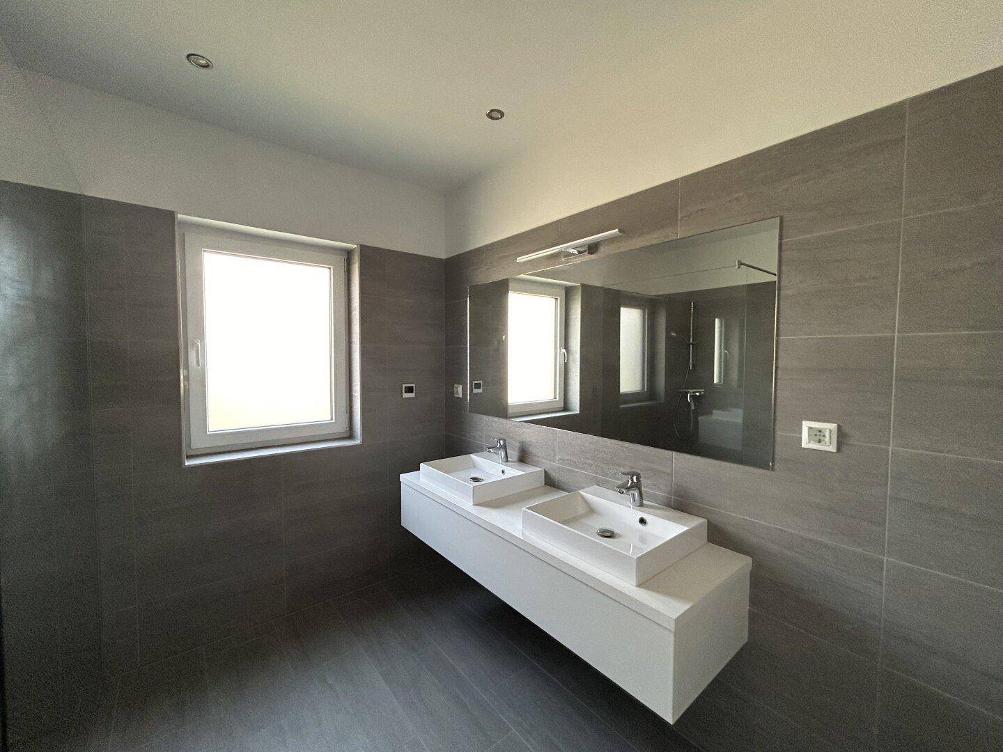 Modernes Badezimmer mit doppelten Waschbecken, großem Spiegel und grauen Fliesen an Wänden und Boden.