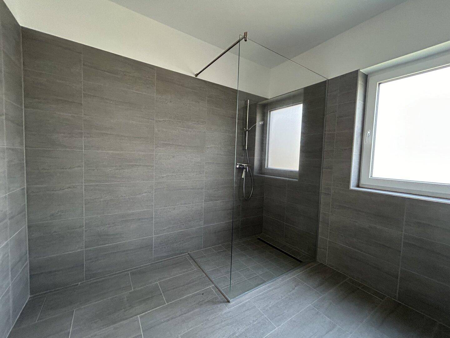 Geräumige begehbare Dusche in einem modernen Badezimmer mit grauen Fliesen und einem Fenster für Tageslicht.