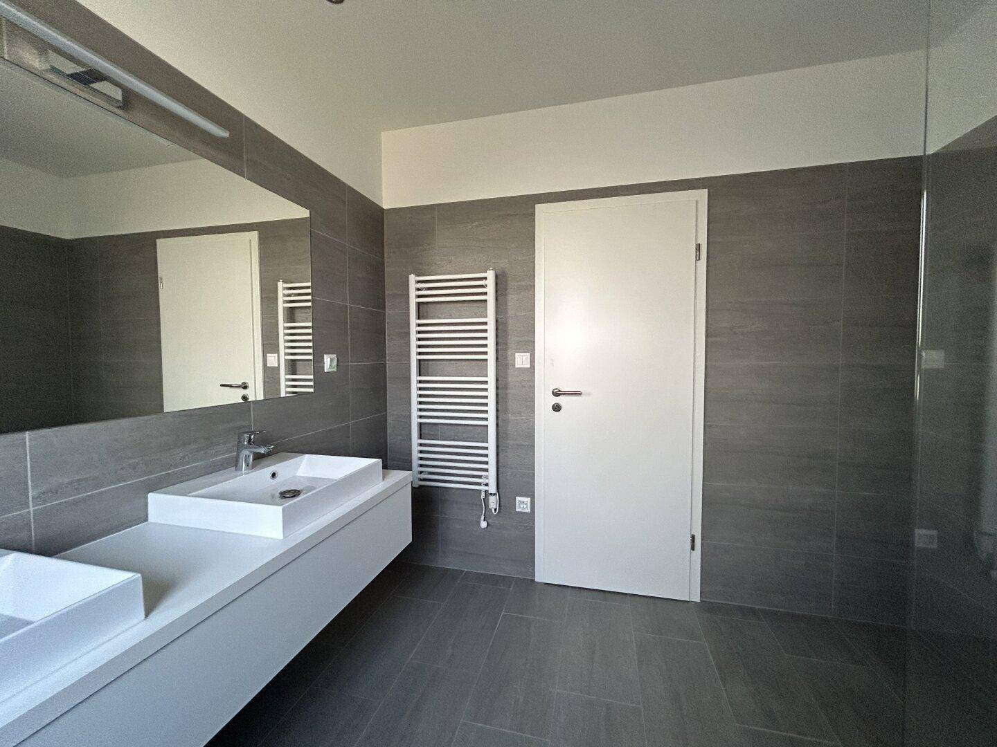 Modernes Badezimmer mit Doppelwaschbecken, beheiztem Handtuchhalter und grauen Fliesen, hell und funktional.