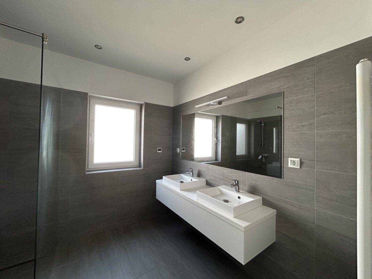 Helles Badezimmer mit zwei Waschbecken, großem Wandspiegel und grauen Fliesen, die eine moderne Atmosphäre schaffen.