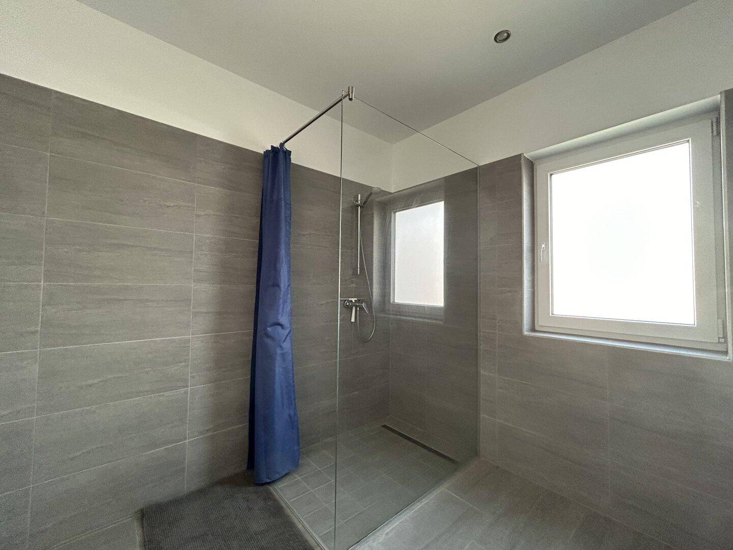 Begehbare Dusche in einem modernen Badezimmer mit grauen Fliesen und einem Fenster, das viel Licht hereinlässt.