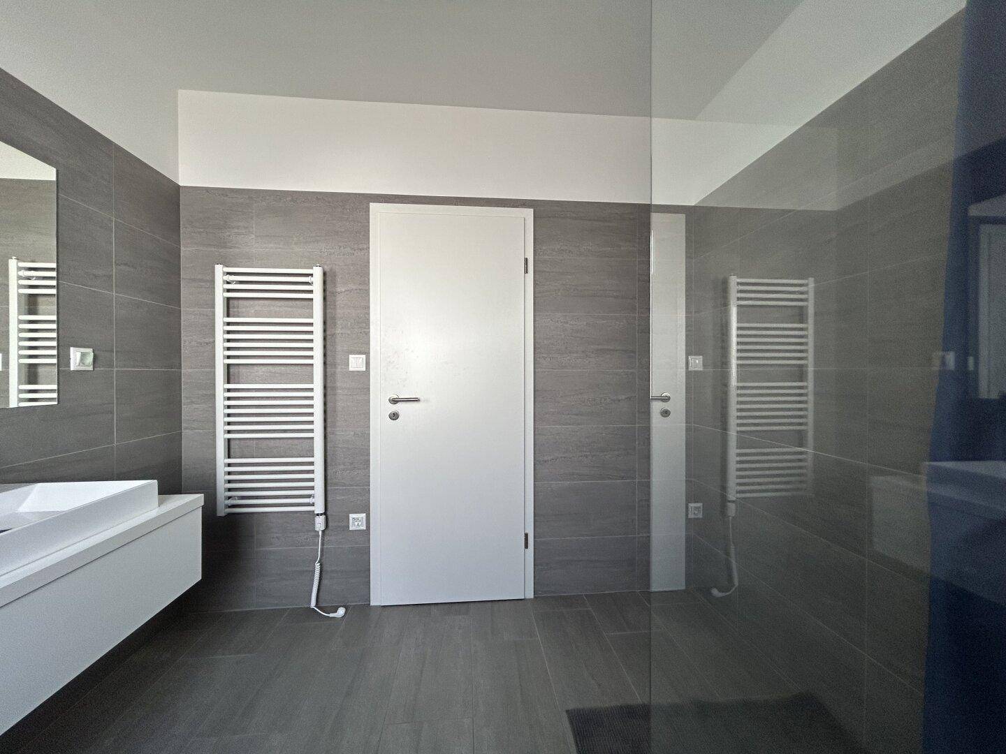 Modernes Badezimmer mit Doppelwaschbecken, beheiztem Handtuchhalter und einer begehbaren Dusche, alles in Grau gehalten.