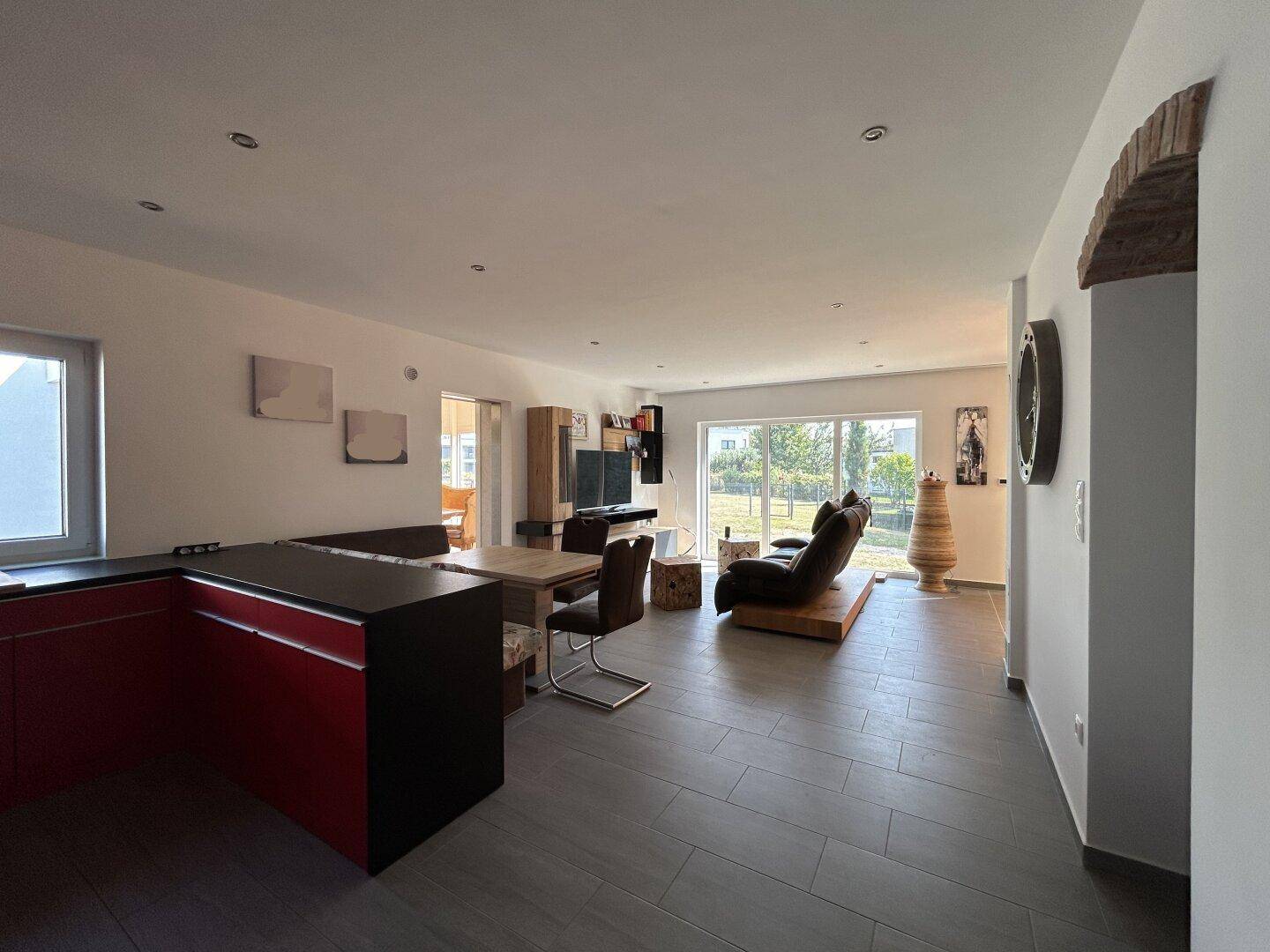Offener Wohn-Essbereich mit roter Küche, Fliesenboden und großem Fenster mit Blick ins Grüne.